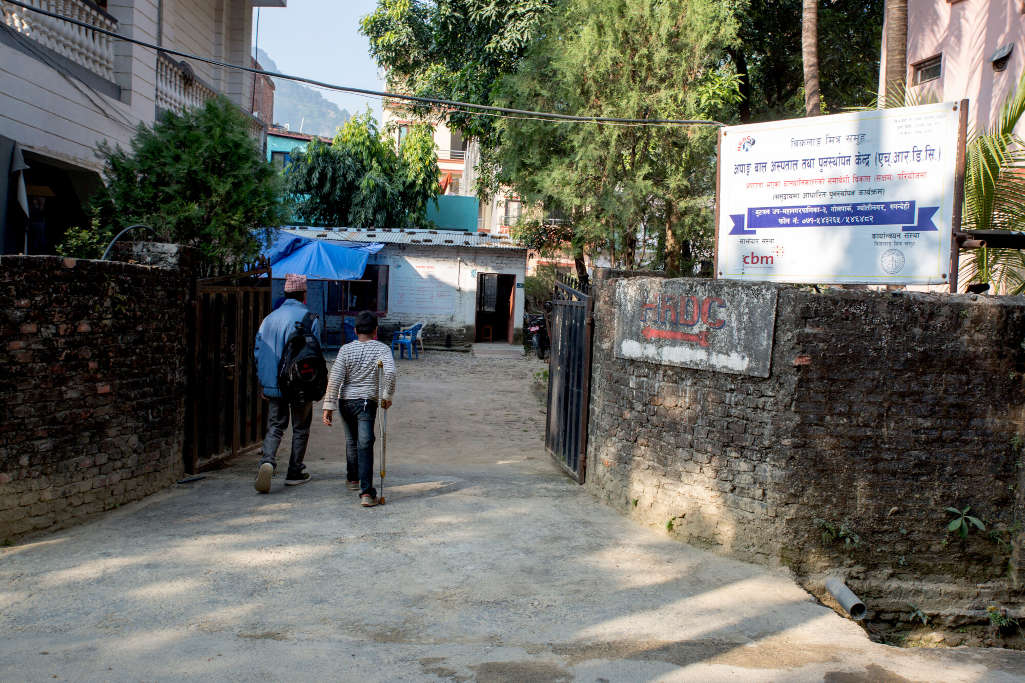 Ingresso della struttura indicata da cartello con scritte nepalesi, papà e bambino di spalle entrano. Papà in giacca azzurra porta lo zaino, bambino con maglia a righe