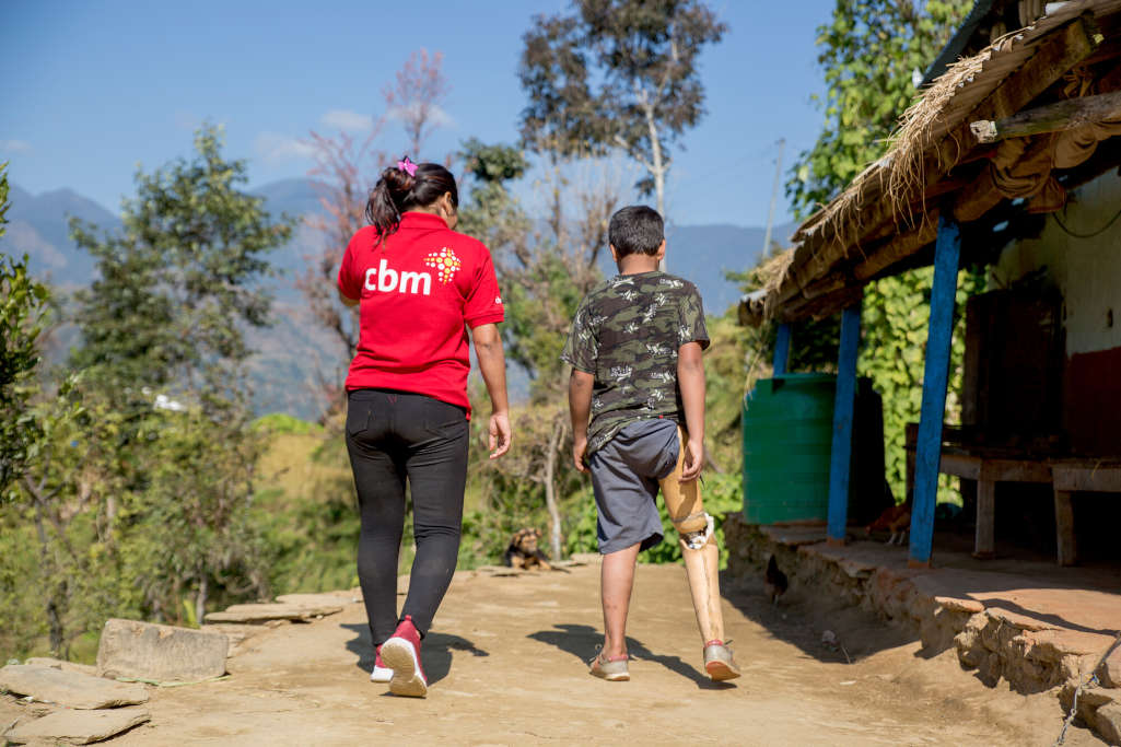 Operatrice accompagna Naresh e controlla come cammina, entrambi di spalle, cortile davanti a casa